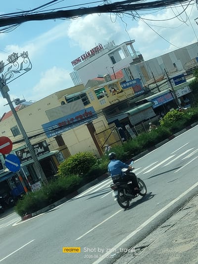 The image captures a street scene in Gò Dầu, Vietnam, on a bright, overcast day. The camera angle is tilted, looking down from a moving vehicle, likely a car or bus.
In the foreground, a person wearing a blue shirt and a black helmet is riding a dark-colored motorcycle down the right side of a paved road. The motorcycle has a license plate that reads "62 B2 - 28". White lane markings are visible on the road. The rider's right leg is bent, and their left leg is positioned over the motorcycle's frame. A black bag is slung across their back.
To the left of the road, there is a sidewalk area with a metal pole supporting traffic signs. One sign is circular with a red border and a horizontal white bar, indicating a prohibition. Below it is a blue circular sign with a white arrow pointing to the left. Next to the pole is a small shop or stall with a blue awning. Further left, a section of green foliage is visible, and behind it, a tall, yellow building with a red-tiled roof. Another building, a clinic identified by the sign "PHÒNG KHÁM BS THÁI" in red lettering, is visible further back, with a modern architectural style and large windows.
The background features a mix of buildings, many with light-colored facades and some with corrugated metal siding. Signs in Vietnamese are present on various buildings, including "sở vật liệu xây dựng" (construction materials) and "mobilefon 5G". Overhead, a dense network of black electrical wires crisscrosses the sky. The sky itself is bright blue with scattered white clouds.
The overall impression is one of a busy urban environment with a blend of commercial establishments and residential structures. The image conveys a sense of everyday life in Gò Dầu.
