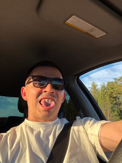 A playfully posed portrait features a man from the chest up, positioned inside a car and looking directly at the camera. He wears dark sunglasses and a white t-shirt. His head is tilted, and he has a humorous expression with his mouth open and tongue slightly extended. Warm, golden sunlight illuminates his face and clothing, suggesting the late afternoon or early evening.
Above the man, the open sunroof of the car reveals a clear blue sky with some scattered clouds, and the lush green tops of coniferous trees, indicating an outdoor setting, likely in a natural or forested area. The interior of the car, including the tan headliner and a sun visor, is visible.
A clear warning label is affixed to the car's sun visor. The text on the label states: "A WARNING DEATH OF SERIOUS INJURY can occur. Children 12 and under can be killed by the air bag. The BACK SEAT is the SAFEST place for children. NEVER put a rear-facing child seat in the front. Sit as far back as possible from the air bag. ALWAYS use SEAT BELTS and CHILD RESTRAINTS. See the Owner's Manual for further information and explanations." The image captures a lighthearted moment, possibly during a car journey in Yus'ki, Russia.