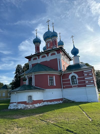 The image shows a red and white domed church with blue onion domes, located in Terpilovo, Russia. The church has multiple domes, each topped with a cross. The main body of the church is painted red with white architectural details, and the lower part is white with red trim. The church is situated on a grassy hill under a bright blue sky with scattered white clouds. To the left of the main church, a smaller yellow building with a green roof is partially visible. Trees are present in the background to the right and left of the church. The foreground consists of a well-maintained green lawn. The lighting suggests it is daytime, possibly late afternoon due to the shadows. There are no people visible in the image.