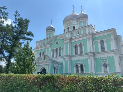 The image shows the Transfiguration Cathedral in Diveyevo, Russia, bathed in sunlight under a clear blue sky. The cathedral is a grand structure with a turquoise facade and white architectural details. It features multiple domes, several of which are topped with golden crosses. Tall, dark green coniferous trees stand to the left of the building, partially obscuring its view. In the foreground, a dense, green hedge runs across the bottom of the frame, with some dried leaves and branches visible within its foliage. The scene is tranquil and showcases the architectural beauty of the cathedral and its surrounding natural elements.