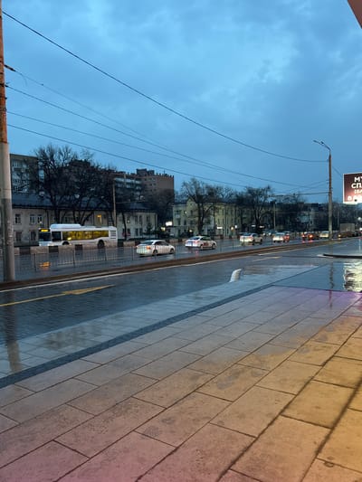 The image depicts a street scene in Ol'ginskaya, Russia during twilight or dusk. The perspective is from an elevated position looking down at an angle. The street is wet, likely from recent rain, with reflections visible on the paved surface. A sidewalk runs along the right side of the frame, made of large rectangular blocks. A bus and several cars are visible in the street. Buildings line the left side of the street, interspersed with bare trees. Overhead wires stretch across the sky, which is a muted blue-gray. A sign is visible in the upper left of the frame, with illuminated text in a Cyrillic script.