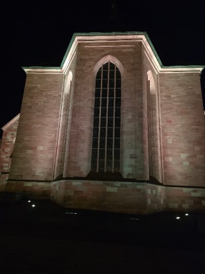 This nighttime photograph captures an exterior view of the Alexanderskirche in Zweibrücken, Germany. The building is constructed from reddish-brown sandstone blocks and is illuminated from below by multiple ground-level spotlights that cast an upward, warm glow onto the facade. The central feature is a tall, narrow, pointed-arch window, divided into numerous dark panes by a grid of mullions and transoms, indicating unlit interior spaces. Architectural elements resembling buttresses or recessed arched panels flank the main window. A distinct green-patinated copper trim or roofline outlines the upper edges of the structure, which presents a multi-faceted or polygonal form consistent with a church apse or chancel. The immediate foreground shows a dark, paved surface, with small, bright ground lights visible at the base of the building. The sky is completely dark, indicating a late-night or very early morning setting. No individuals, vehicles, or discernible actions are present within the frame.