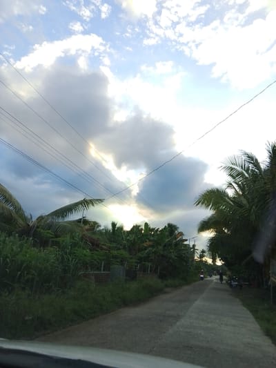 This is an eye-level, medium shot of a rural road in Santa Cruz, Philippines, during what appears to be either sunrise or sunset. The sky is filled with a mix of white and gray clouds, with some areas of bright light suggesting the sun is breaking through. Several power lines are visible crisscrossing the sky. The road itself is paved and curves slightly to the right, disappearing into the distance. On either side of the road, lush green vegetation is abundant, including palm trees and other tropical plants. In the distance, two individuals are riding motorcycles on the road. The overall scene depicts a serene, natural environment with signs of human activity.