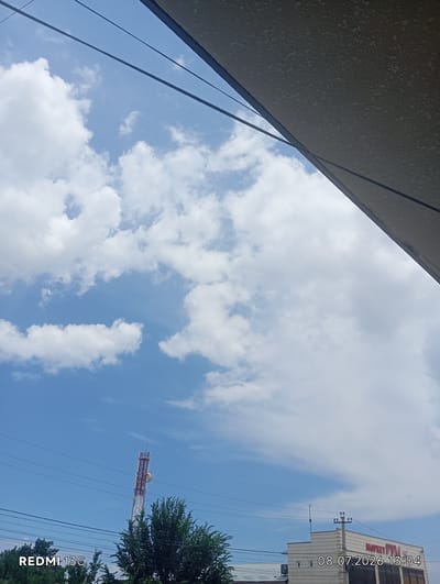 The image depicts a bright, sunny sky with scattered cumulus clouds, characteristic of a clear day. In the foreground, a portion of an overhead structure, possibly a roof or awning, angles across the top of the frame. Power lines are visible crisscrossing the sky, with one prominent tower in the middle left of the image, likely a telecommunications tower, painted red and white. Below the tower, lush green trees are visible. In the bottom right corner of the image, a building is partially visible with text that reads "MARKET PYM" and a date and time stamp "08.07.2026 13:04". The presence of the building with signage and the telecommunications tower suggests an urban or semi-urban environment. The "REDMI 13C" watermark in the bottom left corner indicates the device used to capture the photograph. The overall scene is one of daytime activity under a clear sky, with elements of infrastructure and commerce visible.