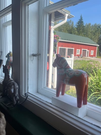 This image depicts an indoor perspective looking out through a white-framed window onto an exterior rural setting in Sipoo, Finland, under clear daylight. The window features white painted wooden sashes and mullions, with a dark green painted sill at its base. A metal window latch is positioned on the central vertical mullion. On the window sill, a reddish-orange Dala horse figurine, adorned with light blue and green patterns, stands on a rectangular white wooden block on the right, facing towards the exterior. To the left, a dark grey or black ornate horse figurine, appearing to be crafted from metal or resin, is positioned on a dark, curved metal stand, facing left. A third, light green and colorful Dala horse is partially visible and out of focus behind the dark figurine. The exterior background features a prominent red wooden barn or outbuilding with a dark green pitched roof. This structure includes two large, dark garage-style doors with white trim on its left side and multiple white-framed windows on its right. A white downspout is visible on the building's right exterior. The immediate outdoor ground comprises green grass and tall green vegetation. Beyond the red building, dense green coniferous trees are visible under a clear blue sky.