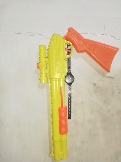 The scene depicts three objects positioned against a light-colored, textured wall exhibiting multiple cracks. The specific location within Umsobomvu Local Municipality, South Africa, is not identifiable from the image.
The primary object is a large, yellow plastic toy gun, designed to resemble a rifle or shotgun, oriented diagonally with its stock pointing towards the upper right and its barrel towards the lower left. It features an integrated yellow scope on the top and an orange pump-action foregrip towards the lower barrel. Adjacent to the upper right section of this yellow toy gun, an orange plastic toy gun stock is positioned, appearing separate and leaning against the wall, slightly overlapping the yellow toy gun. Between the trigger guard area of the yellow toy gun and the wall, a silver-colored wristwatch with a black face and a metal mesh strap hangs vertically. The watch face prominently displays a "ck" logo, indicative of the Calvin Klein brand. No individuals are present, and no actions beyond the static arrangement of objects are depicted.