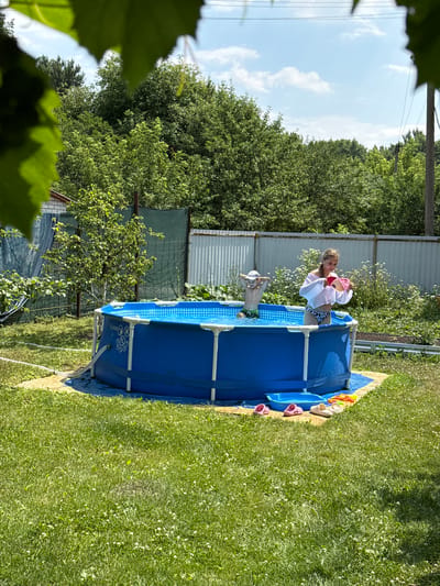 In a backyard in Nizhne-Chufichevo, Russia, on a sunny day, two children are playing in a blue inflatable swimming pool. A boy, wearing a white cap and dark swim trunks, stands in the water. A girl, wearing a white semi-sheer shirt and patterned bikini bottoms, stands in the pool holding a pink toy. The pool is on a patch of grass, with a blue tarp underneath. Outside the pool, on the grass, are several pairs of children's shoes and a small blue water toy. The background is filled with lush green trees and a metal fence. A hose is visible leading from the pool. The scene suggests a recreational activity during warm weather.