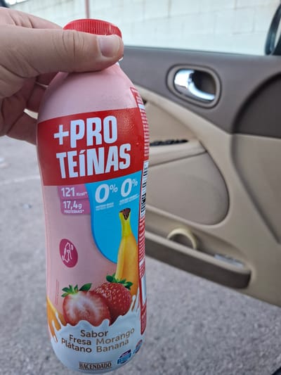 A hand holds a bottle of "+" Pro Teinas protein shake, strawberry and banana flavor, branded "Hacendado." The bottle is predominantly pink with red and white lettering and images of a banana and strawberries. The label states "0% Materia Grasa/Materia Gorda" and "0% Azucares Anadidos/Acucares Adicionados." The background shows the interior of a vehicle with a beige door panel and silver door handle, and outside the car, a gray asphalt surface. It is unlikely to be in Quart de Poblet, Spain, as the description is based solely on the details in the provided image.