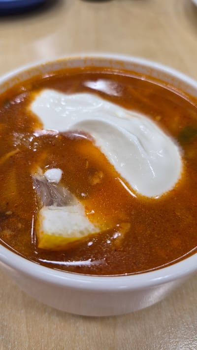 A close-up, overhead shot shows a white bowl filled with a hearty, reddish-orange soup. A dollop of thick, white sour cream is swirled on top of the soup. A piece of lemon or white cheese, possibly with a clear plastic wrapper, is partially submerged in the soup. The soup appears to have small pieces of ingredients within it. The bowl rests on a light brown, textured surface, likely a wooden table. The background is softly blurred, with a hint of a blue object at the top. The image captures the soup in a restaurant or home setting, with no specific geographic indicators of Preiļi, Latvia.