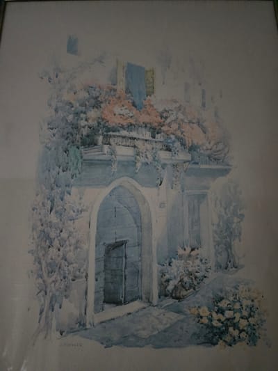 A watercolor painting depicts a doorway in a building, surrounded by lush greenery and flowers. The architectural style suggests an older European setting. The doorway is arched and leads into a dark interior. Above the doorway, a balcony is adorned with plants and flowers in shades of pink and white. The overall impression is one of a tranquil and picturesque scene. The artist's signature, "J. KOEHLER," is visible in the lower left corner. This painting is believed to be a depiction of a location in the city of Mutzig, France.