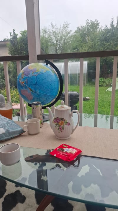 This is a still life photograph taken outdoors on a covered porch or deck overlooking a yard in the city of Орбита Снт (терпигорьево), Russia. The scene is set on a glass-topped table with a cowhide-patterned rug underneath.

On the table, there is a globe, a white ceramic teapot with a floral pattern, a can of beer, a white mug, a bag of chips, and a red package of Ritter Sport marzipan chocolate. The globe is positioned on the left side of the frame, showing Asia and Australia. The teapot, adorned with pink and yellow roses, is in the center. The beer can, partially obscured, is to the left of the teapot. The mug is in front of the can, and the bag of chips is to the left of the mug. The chocolate bar is on the right side of the table, partially overlapping the edge.

Through the railing and glass of the porch, a green lawn is visible with a greenhouse and a barbecue grill in the background. The sky is overcast, suggesting a cloudy day. The overall impression is one of a relaxed, domestic setting, possibly during a break or a casual gathering. There are no people visible in the image.