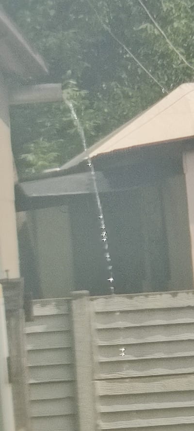 A vertical, narrow stream of water cascades down what appears to be a window or transparent surface. The water droplets are illuminated by an unseen light source, creating a chain of bright spots. The background is out of focus and shows greenery, likely trees and bushes, suggesting an outdoor environment. A portion of a building with a light-colored roof is visible. In the foreground, a slatted fence-like structure is present. The scene is consistent with a water stream on a window in Shelestove, Ukraine, possibly from a sprinkler or a leaky gutter.