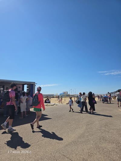 The media file depicts a sunny day at a beachside promenade in Le Havre, France. The clear blue sky dominates the upper portion of the image, with a few wispy clouds. The ground is a paved surface with visible shadows cast by people, indicating a bright, sunny time of day.
The scene is populated by numerous people walking along the promenade, some casually strolling, others in groups. A food stand or kiosk is visible, with people queuing or interacting with it. The beach itself is in the background, with sand and beachgoers visible.
Notable details include a person in the foreground wearing a bright red jacket and green shorts, with a backpack. Another person in a pink top is also prominent in the foreground. Several individuals are wearing masks, possibly reflecting a past or ongoing health precaution. The overall atmosphere appears relaxed and casual, typical of a seaside outing.
There is text visible in the lower right corner of the image: "17 юни 2026 г.", which translates to "June 17, 2026". This suggests the image was taken on that date. The event taking place is a public gathering on a promenade adjacent to a beach.