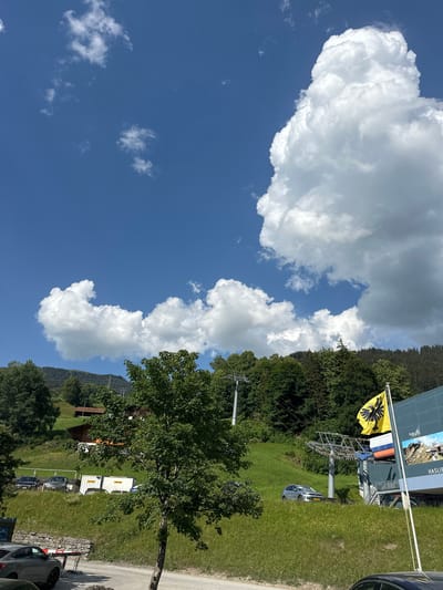 The image depicts a mountain landscape in Hasliberg Reuti, Switzerland. In the foreground, a dirt path runs horizontally, with a stone wall on the left and a dark colored car parked on the right. A tree with green foliage stands prominently in the middle ground, obscuring part of a grassy slope behind it. Several cars are parked on the grassy slope, and two white shipping containers are visible. Further up the slope, a ski lift with its towers and cables stretches across the frame, ascending towards a densely forested mountain range. A yellow flag with a black eagle emblem flies on a pole to the right of the ski lift, next to a building with a large advertisement. The sky is a vibrant blue, dotted with large, white, cumulus clouds. The overall scene is one of a sunny day in a mountainous, recreational area.