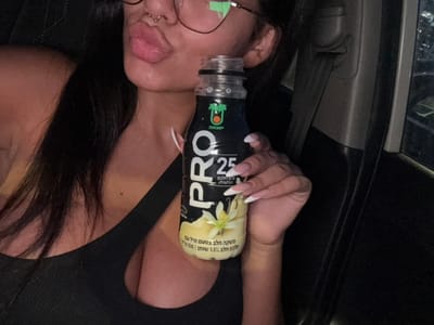 The image shows a young woman holding a bottle of "PRO" coconut milk, a brand sold in Israel. The woman is seated in the driver's seat of a car. She is wearing a black tank top and has long dark hair. She is holding the bottle with her right hand. The bottle is partially visible and the text on the label is in Hebrew. The woman is looking at the camera and is smiling. The background is dark. The image was taken in Ramla, Israel, at the coordinates 31.931, 34.853.