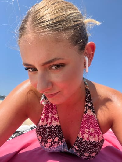 The image displays a female individual situated on a sandy beach in Ribamontán al Mar, Spain. The subject, with blonde hair pulled back, is observed in a prone or leaning posture, looking directly towards the camera. Her skin exhibits a tanned tone and a visible sheen. A white Apple AirPod is present in her right ear. She is wearing a halter-neck bikini top with a multi-color pattern consisting of pink, black, brown, and light blue sections. The individual is positioned on a pink towel or mat. A portion of another patterned textile, displaying black, white, and gray tones, is partially visible to the left, beneath the pink towel. The background is a clear, unclouded blue sky, with a faint, indistinct landmass or coastal feature visible on the far left horizon.