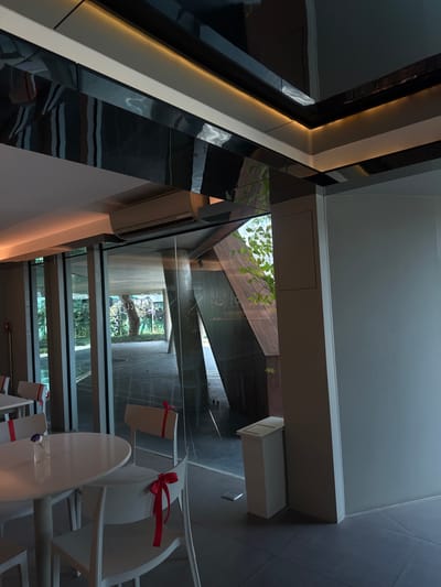 An architectural interior showcases a modern design. A black, white, and gray structure with reflective surfaces defines the left side. A glass corridor reveals a concrete structure and natural greenery. White chairs, each decorated with a red bow, are arranged near a table, suggesting a dining or gathering space. The composition is sharp and angular.