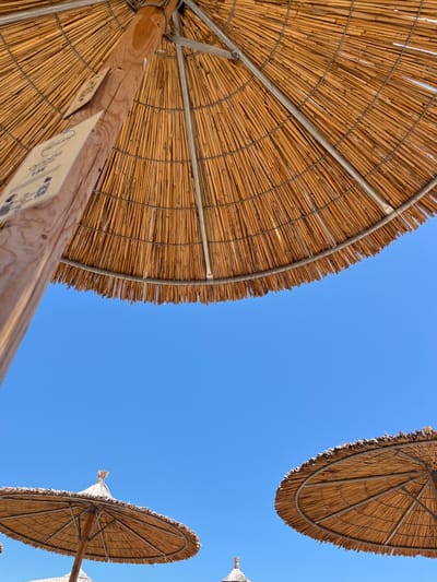 The image depicts a clear blue sky with two thatched beach umbrellas. The main umbrella in the foreground is viewed from below, showing its woven straw canopy supported by a wooden pole and metal struts. A sign is attached to the pole. In the background, another thatched umbrella is partially visible. The scene is likely a beach in Νέα Ηρακλείτσα, Greece, given the context.