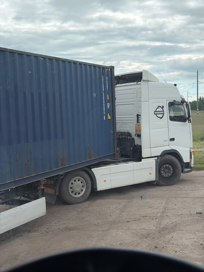 A white Volvo semi-truck, model 400, is parked on a dirt road next to a large, dark blue shipping container. The truck has the Volvo logo on its door. The shipping container is positioned at an angle on the truck's trailer. The environment is outdoors with a cloudy sky above and a grassy area with trees and power lines visible in the background. The scene appears to be in a rural or industrial area. The exact location is Zarech'e, Russia.