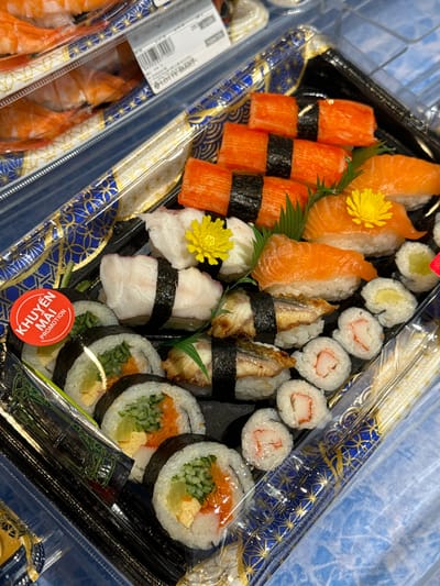 The image shows a take-out sushi container with various types of sushi and sashimi. The container is filled with a variety of sushi rolls, nigiri, and what appears to be imitation crab sticks. Some pieces have garnishes like yellow flowers and green leaves. There's also a red circular sticker with Vietnamese text that says "KHUYEN MAI PROMOTION". The container is placed on a surface with a blue and gold patterned design, possibly a tray or table. The overall presentation suggests a food product ready for sale or consumption. The image does not provide any specific visual information to pinpoint its location within Phu Thuy, Vietnam.