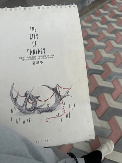 The image shows a notebook resting on someone's lap outdoors. The notebook cover has the title "THE CITY OF FANTASY" printed on it, along with other text in both English and Chinese. The illustration on the cover features a drawing of what appears to be a monster or dragon-like creature with a jagged, red-lined interior. In the background, there are patterned brick paving stones. The lower right corner of the photo includes the edge of a light-colored garment and a glimpse of a white sock and a black shoe. The text "SHOWYOURWAY" is also visible on the right side of the notebook cover. The location, Agarak, Armenia, is not deducible from the image alone.