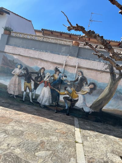 A large mural depicting eight figures in 18th-century attire engaged in a circular dance dominates the wall of a building in Piedrahíta, Spain. The figures, four men and four women, are rendered in a style reminiscent of Goya's tapestry cartoons, with vibrant clothing including feathered hats, flowing dresses, breeches, and jackets. A painted landscape with rolling hills and a light blue sky serves as the background for the dancers. Two white vertical pipes are affixed to the wall, passing through the center of the mural and partially obscuring two figures.
Above the mural, the building wall features a grey-blue horizontal band, topped by a decorative white railing with a geometric pattern, suggesting a balcony or terrace. A light-colored building with a sloped tile roof is visible behind the railing, with a terracotta pot containing a small green plant situated on the railing to the left. A television antenna is mounted on a rooftop in the upper right section of the image.
In the foreground, the ground is paved with irregular, light-colored flagstones, displaying a distinct straight line of lighter pavers running from the lower center towards the mid-right. A bare-branched, gnarled tree with an unpainted trunk and branches extends from the upper right, partially obscuring the top right portion of the mural. The base of this real tree appears to blend seamlessly with a painted tree trunk on the mural. The scene is illuminated by direct sunlight, casting distinct shadows on the ground and parts of the mural from the top left.