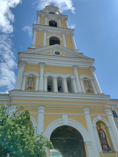 This is a low-angle, full-shot photograph of the bell tower of the Trinity Cathedral in Diveyevo, Russia, on a clear day. The tower is painted yellow with white architectural elements, including columns, arches, and decorative moldings. The sky is a vibrant blue with scattered white clouds.
The bell tower is tall and slender, tapering towards the top. It features multiple levels with arched openings. On the lower levels, there are mosaic icons of religious figures embedded in niches. One mosaic depicts an angelic figure holding a staff, and another shows a robed figure with a halo. The largest arch at the base of the tower appears to lead into an interior space decorated with further mosaics.
A lush green tree with dense foliage partially obscures the bottom left of the bell tower. Several small, dark birds are visible in the sky, appearing as specks against the blue. The overall impression is one of grandeur and religious significance.