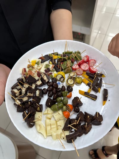 A white plate filled with assorted bite-sized snacks, each individually speared with a toothpick, is held by a person wearing a black shirt. The plate contains dark chocolate pieces studded with nuts, white chocolate pieces, dark chocolate rolls with white centers, dark chocolate pieces with yellow fruit, green olives, small red candies, and some dried fruit. The snacks are arranged somewhat haphazardly on the plate. The background suggests an indoor setting with tiled walls and flooring. The person holding the plate has their arms and torso visible, and a partial view of another person's hand and foot wearing sandals is visible on the right side of the image. The lighting is bright. The location in Phu Thuy, Vietnam is not discernible from the image.