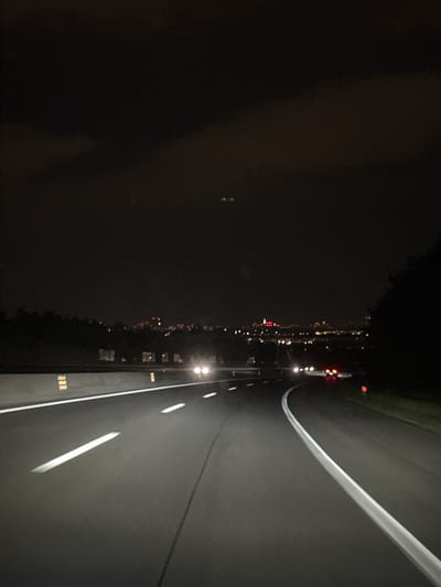 The image depicts an aerial view at night of a road, likely located on the outskirts of or just outside Brunn am Gebirge, Austria, judging by the scattered lights that could be from the town. The road is dark grey or black with white painted lines indicating lanes. There are also reflectors or markers along the side of the road. The background is predominantly dark, suggesting undeveloped or rural areas beside the town. The overall impression is that this is a road winding into or out of Brunn am Gebirge at night.