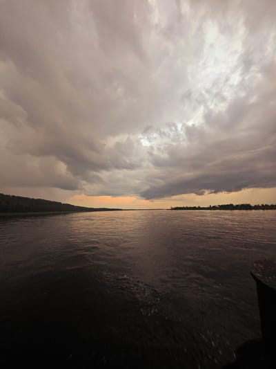 The image depicts a wide, dark river flowing through a landscape under a cloudy, overcast sky. The sky is filled with heavy, grey clouds, with a sliver of pale orange light visible near the horizon, suggesting either dawn or dusk. The left bank of the river features a dense, dark green treeline. The water's surface is rippled, with some white foam or spray visible in the foreground, indicating movement. In the far right foreground, a dark, rounded object, possibly a boat fender or mooring bollard, is partially visible, suggesting the perspective is from a vessel on the water. The overall atmosphere is somber and subdued. The location is specified as the city of Kazachinskoe, Russia, likely referring to the Angara River or a similar large waterway in the region. There are no people or animals visible in the image.