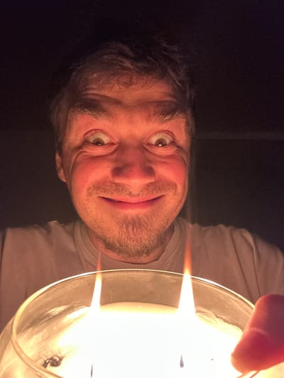 A close-up, selfie-style image shows a Caucasian male holding a large, lit candle in a dimly lit environment. The man is positioned in the center of the frame, with his face illuminated by the candle flame. He has light brown hair, parted slightly to his right, and appears to be in his late 20s or early 30s. His eyes are wide open and looking directly at the camera, with his left eye appearing slightly dilated. He has a wide, exaggerated smile, revealing his teeth, and a light beard and mustache. His skin is flushed red from the proximity to the candle. He is wearing a light-colored t-shirt. The candle, with two visible wicks, is in the foreground, taking up the bottom third of the image. The background is dark and out of focus, suggesting a private indoor setting. The lighting is dramatic, with the warm glow of the candle casting strong highlights and shadows on the man's face. The image is a selfie, indicated by the perspective and the man holding the camera. The specific location in the city of Рассоха, Russia, is not discernible from the image itself.