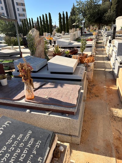 The image depicts a cemetery in Gedera, Israel, under a clear blue sky. The scene is dominated by rows of graves with various types of headstones, some made of granite and others of a lighter stone. Several graves are adorned with plants, flowers, and decorative stones. In the foreground, a vase with dried flowers sits on a grave. Behind the graves, a tall, modern building stands, contrasting with the older, more traditional cemetery setting. The ground is a mixture of dirt and paved paths. The overall impression is one of a well-maintained, active cemetery.