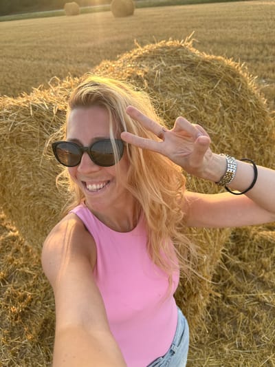 A young woman with blonde hair and sunglasses is taking a selfie in a field of hay bales. She is wearing a pink top and denim shorts. The sun is setting, casting a warm glow on the scene. The woman is smiling and holding up two fingers in a peace sign. The background shows several large hay bales and a distant hill. The location is identified as Trouville-Alliquerville, France.