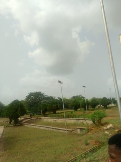 The image depicts an outdoor, landscaped park area in Adzho, Nigeria, under an overcast sky filled with dense grey clouds. The foreground consists of unkempt green grass, transitioning into a multi-tiered section with concrete retaining walls and pathways. Numerous trees and shrubs are distributed throughout the scene; some are large, mature trees with full canopies, while others are smaller, conical-shaped shrubs. Several tall, slender poles, equipped with what appear to be solar-powered light fixtures or surveillance equipment, are spaced across the park.

In the mid-ground, two figures are visible on a concrete pathway; one, wearing dark attire, is positioned near a large tree, while the other is further along the path. A third distant figure, wearing a light blue or green shirt, is discernible amidst the shrubs. In the immediate bottom right corner, the right side of a human face, featuring a dark complexion and an ear, is partially visible, indicating a close foreground presence or the photographer's perspective. The overall scene suggests a public park or green space with varying levels of maintenance.