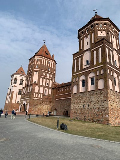 An exterior long shot depicts the Mir Castle Complex in Мірскі сельскі Савет, Belarus, under a partly cloudy sky. The prominent structure is a multi-towered castle constructed primarily of red brick with sections of lighter pink/beige stucco or plaster detailing. The largest tower on the right features a stone and brick base, transitioning to predominantly brick with vertical stucco strips and arched window surrounds, culminating in a pyramidal reddish-brown tiled roof topped with a weather vane. A smaller, lighter-colored tower to the left exhibits a similar tiled roof and white stucco facade sections with dark-framed windows. Connecting brick walls are visible between the towers. In the foreground, a gray paved pathway curves from left to right, bordered by a strip of sparse green grass. Approximately ten to twelve individuals, dressed in casual attire, are distributed along the pathway and near the castle walls, appearing to be walking or observing the structure. Ground-level bollards or lights are positioned along the edge of the grass, and a decorative element resembling an antique cannon wheel is visible on the grass to the right.