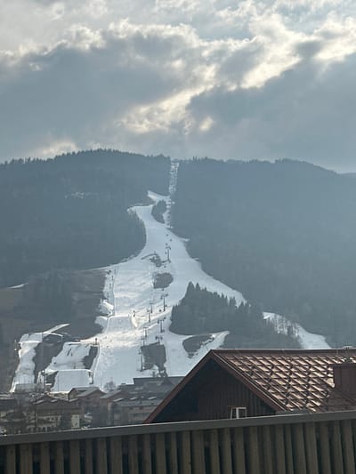 The image displays a ski resort in Sankt Georgen am Kreischberg, Austria, under an overcast sky featuring grey and white clouds. A large, forested mountain occupies the background, with a distinct, wide ski slope covered in artificial white snow descending its face. Multiple ski lift towers with cables are visible ascending the slope. At the base of the mountain and ski slope, an alpine village comprising several buildings with gabled roofs and light-colored facades is discernible. The immediate foreground shows the dark red, tiled gabled roof of a building and a horizontal railing composed of vertical slats, partially obscuring the view of the village. No individuals are clearly visible on the ski slopes, ski lifts, or within the village.