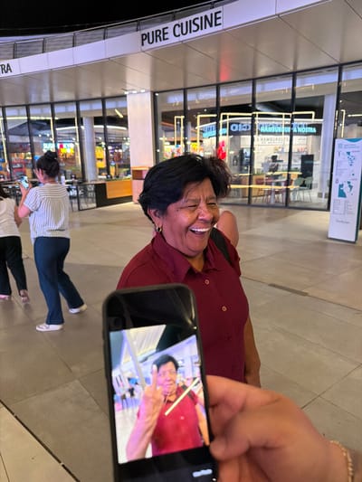 The image depicts an outdoor, public walkway, possibly a mall or entertainment district, with a prominent sign reading "PURE CUISINE" overhead. In the foreground, a person's hand holds a smartphone, displaying a photo of a man making a peace sign. To the right of the phone, an elderly woman with dark hair and a maroon shirt is laughing with her eyes closed. Behind her, two other individuals are visible; one woman in a striped shirt and dark pants is walking away, and another woman in a white shirt is partially visible. The background features storefronts with illuminated signage, suggesting commercial establishments. The lighting indicates it is nighttime or late evening. The scene appears to be in the city of Alcorcón, Spain, given the context of the provided hint, though this cannot be definitively confirmed from visual cues alone.