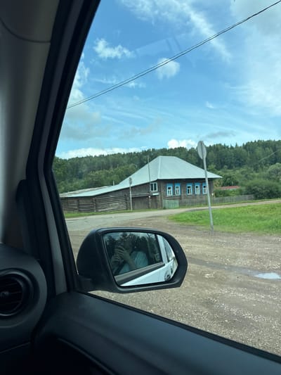 The image, taken from inside a vehicle, shows a view through the passenger side window and the side-view mirror. The scene is a rural area in or near Mostovaya, Russia, featuring a dirt road, grassy areas, and a dense forest in the background. A wooden house with a light-colored metal roof and bright blue window frames is visible. Adjacent to it is a longer wooden structure, also with a metal roof. A circular road sign is positioned near the house. The side-view mirror reflects the interior of the vehicle, showing a person holding a phone, possibly taking a selfie. The sky is blue with scattered white clouds.