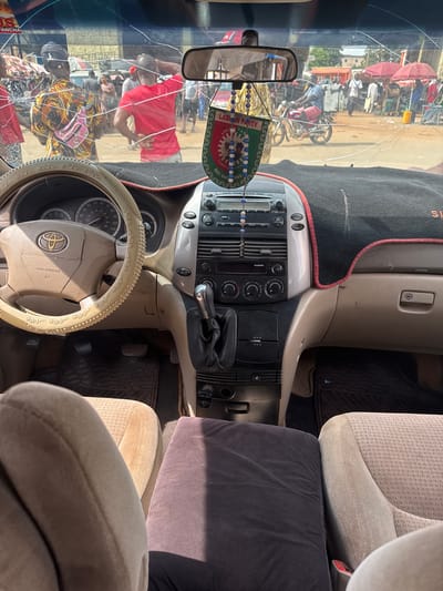 The image is taken from inside a vehicle, looking out through a cracked windshield at a bustling street scene in Orji, Nigeria. The dashboard and steering wheel are visible in the foreground, with a beige interior. A steering wheel cover with Arabic script is present. A car mat is visible on the floor.

Outside the vehicle, a group of people are gathered. One person in the foreground to the left wears a colorful patterned shirt and a pink fanny pack. Another person in the center is wearing a red t-shirt. There are market stalls with red umbrellas and various goods displayed. A motorcycle is visible in the middle ground. The environment appears to be a busy outdoor market or street. A "LABOUR PARTY" banner is hanging from the rearview mirror.