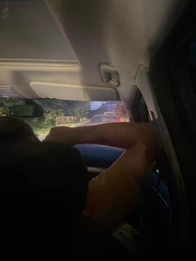 The image shows a person's legs and torso inside a car, with the car's interior visible in the foreground. The person is wearing tan shorts. Outside the car, through the windshield, trees and a red ladder are visible. The lighting suggests it is dusk or nighttime, with artificial lights from what appears to be a street or another vehicle reflecting on the car's interior and windshield. The scene appears to be in a somewhat rural or suburban setting, given the trees and ladder. The angle of the photo is unusual, pointing upwards and outwards from inside the car, making it difficult to discern the exact activity taking place. No text is visible in the image. The location context provided is Beloci, Moldova.