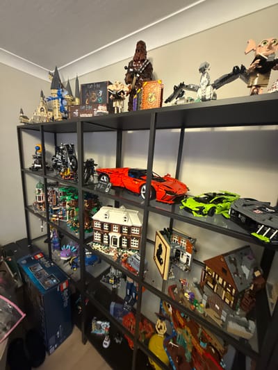 This media file depicts a collection of Lego sets displayed on black metal shelves. The items are predominantly Lego replicas of vehicles, buildings, and characters, many of which appear to be from the Harry Potter and automotive franchises.
On the top shelf, a Lego figure resembling Dobby from Harry Potter is visible, alongside what looks like a Lego Millennium Falcon. The next shelf down features several Lego cars in vibrant colors, including a bright green sports car and a red supercar. Further down, various Lego buildings and dioramas are arranged, including a detailed Lego house with multiple windows and a Lego Hogwarts castle. There are also Lego figures and smaller Lego creations interspersed throughout the display.
The lighting suggests an indoor setting, likely a room in a home, with the shelves illuminated by overhead light. The overall impression is one of a curated collection, meticulously arranged for display. Visible text on some of the Lego boxes and instruction manuals includes "LEGO" and "Harry Potter." The setting is implied to be Newcastle-under-Lyme, United Kingdom, based on the provided location context. There are no people actively engaged in an activity; the focus is on the Lego models themselves. The image is taken from a slightly elevated angle, looking down at the shelves.