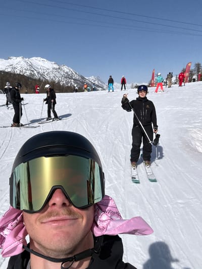This image captures a wide-angle, low-angle selfie perspective from a ski slope in Saint-Chaffrey, France, under clear sunny conditions. In the immediate foreground, the lower face and upper torso of a male individual are visible. He wears a black ski helmet with large, reflective gold-tinted goggles (Oakley Prizm branding visible) and a light brown mustache. A pink bandana with a white pattern is visible under his helmet straps, covering his ears. He displays a faint smile and holds the camera.

Directly behind him and slightly to the right, a younger individual stands upright on skis, facing the camera. This person wears a dark ski jacket with an Arc'teryx logo, dark ski pants, a blue helmet, and goggles. They hold two ski poles, with their right hand raised, extending an index finger upwards. Their skis have noticeable light green sections on the tips and tails.

The midground and background feature a wide, groomed, white snowfield sloping gently upwards. Numerous other individuals (approximately 15-20) are scattered across the slope, either standing or actively skiing. Their attire includes various bright colors such as red, light blue, and dark blue, with some also in black. One skier to the far left mid-ground is notably dressed in a dark suit jacket over ski pants. In the distant background, towering snow-capped mountain peaks rise under a clear blue sky, with dark green coniferous trees sparsely populating their lower slopes. Overhead, thin dark lines representing ski lift cables traverse the upper portion of the sky. Red and yellow flags or banners are visible on the right side of the background slope. Shadows cast on the snow indicate strong direct sunlight.
