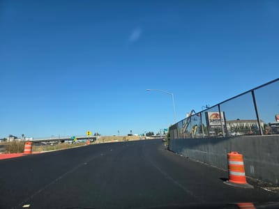 This media file depicts a road under construction in West Sacramento, California, likely during daylight hours under a clear blue sky. The scene is dominated by a freshly paved asphalt road curving to the right, with construction cones strategically placed along the edge. In the background, an elevated highway overpass is visible, with vehicles driving on it. Construction equipment, including cranes and excavators, are present on the far side of the road, hinting at ongoing work. A chain-link fence separates the construction area from a building complex with multiple stories. A sign for "SUNSTATE Equipment Rental" is also visible, further reinforcing the construction theme. The overall impression is one of active infrastructure development, with no visible people or overt signs of emotion, emphasizing the functional and developmental nature of the scene.