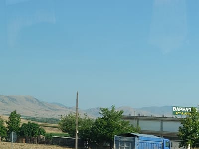 The image captures a daytime outdoor scene under a clear blue sky. In the foreground, a blue truck is partially visible on the right side. A wooden pole stands vertically in the middle of the frame, with a wire extending from it diagonally upwards to the right. Behind the pole and truck, a row of green trees is prominent. In the mid-ground, a building with several large windows is situated, and above it, a green sign displays white lettering that appears to be in Greek: "ΒΑΡΕΛΟ !!", and below that, "Α θει Α λιο". In the background, rolling hills and mountains with sparse vegetation stretch across the horizon, suggesting a rural or semi-rural environment. The overall lighting indicates it is a sunny day.