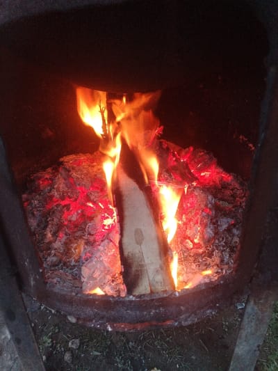 An actively burning fire is depicted within a dark, circular metal containment structure, possibly a fire pit or the base of an outdoor oven. Bright orange and yellow flames rise from several pieces of wood, including a prominent vertical log, which is partially consumed by fire. Below and around the burning wood, a dense bed of red-hot embers and white-grey ash is visible, indicating sustained combustion. The fire serves as the primary light source, illuminating the interior of the structure. The ground immediately surrounding the metal container consists of dark soil with sparse, short green vegetation. No individuals, animals, or specific actions are discernible. The scene is located in Nurli zamin, Uzbekistan.