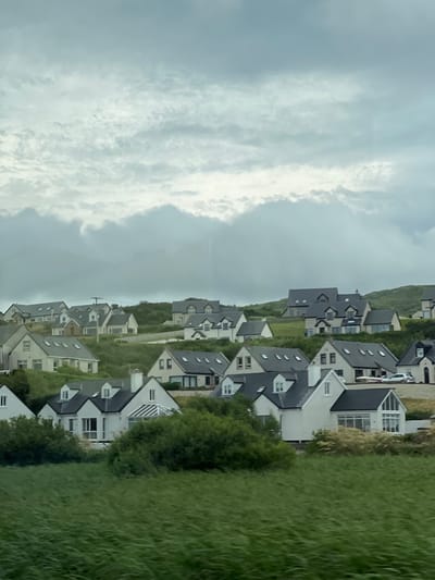 This is a daytime, eye-level, medium shot photograph of a residential development on a hillside in Dunfanaghy, Ireland. The sky is overcast with heavy, textured clouds. The houses are modern, with white facades and dark grey pitched roofs, many featuring dormer windows and large windows. They are clustered together, situated on a grassy, undulating slope. The foreground is dominated by a field of tall, green grass that appears to be windblown, creating a blurred, streaky effect. The houses are arranged in rows, with some appearing larger and more detached than others. The overall impression is one of a relatively new housing estate, possibly a holiday or second-home development, set against a natural, somewhat rugged landscape. There are no people visible in the image, and the scene appears to be still and quiet. The photograph was likely taken from a moving vehicle, given the motion blur in the foreground grass.
