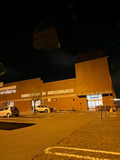 This nighttime image depicts the exterior of the Esselunga di Buccinasco supermarket in Buccinasco, Italy. The primary subject is a large, multi-story commercial building with a brick facade, featuring the illuminated white text "ESSELUNGA DI BUCCINASCO" across its upper section. A central pedestrian entrance is visible, where two individuals are standing. To the right, a wider service bay entrance is marked with the Italian word "ENTRATA" (Entrance).
The foreground shows an asphalt parking lot with clearly painted white markings and a distinct section of cobblestone pavement on the right. A white five-door hatchback car is parked on the left side of the frame, and a light-colored SUV is positioned closer to the building's central entrance. A bare, deciduous tree stands within a planted island in the parking area. Various traffic signs, including blue parking signs marked 'P A' and 'P B', and bollards are present. A cluster of metal shopping carts is visible near the rightmost "ENTRATA" bay. The scene is illuminated by warm-toned artificial lighting, creating shadows on the ground under a dark, clear night sky. A reflection or internal structure from the capturing device is partially visible at the top of the frame.