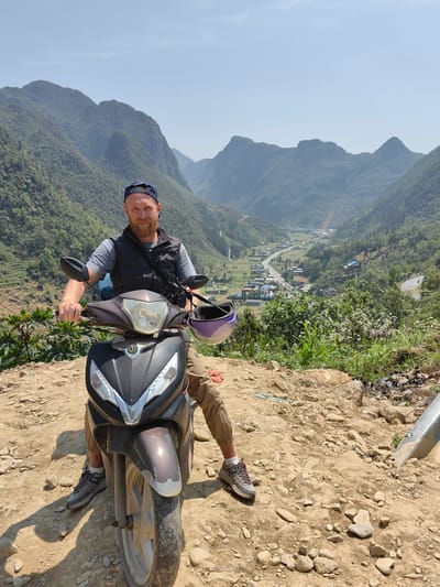 A man sits on a moped on a dirt hill overlooking a valley in Phố Bảng Commune, Vietnam. The man has a beard, wears a baseball cap, a grey t-shirt, and a black vest. He is looking towards the camera. A purple helmet is strapped to the moped. Behind the man and the moped is a valley with a road running through it, surrounded by mountains. The foreground is a rocky, dirt hill with some vegetation. A metal pole is visible to the right of the moped.