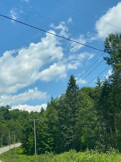 The image depicts a daytime scene in Lac-Simon, Canada, characterized by a bright blue sky filled with fluffy white clouds. Several power lines stretch diagonally across the frame, intersecting with a dense forest of green trees that covers a hillside. In the foreground, lush green foliage and some smaller bushes are visible. To the left of the frame, a paved road curves into the distance, with a speed limit sign indicating "70". Two wooden utility poles stand along the side of the road, supporting the power lines. The overall environment suggests a rural or semi-rural setting with natural beauty and infrastructure. There are no people or animals visible in the image.