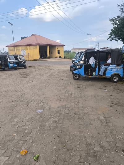 The image depicts a daytime scene at a tricycle (keke Napep) park or station in Obiaruku, Nigeria, situated near a university study center. The ground is a blend of uneven dirt and interlocking pavers, with some scattered litter including leaves and plastic debris.

Several blue and white tricycles are present. On the right, a blue tricycle is prominently parked with three individuals. A man in a white shirt is in the driver's seat, while a woman in a white shirt and blue jeans sits in the back, looking towards the camera with a relaxed expression. Another person is partially visible behind the driver. The side of this tricycle features text, including "DELTASTATE UNIVERSITY ABRAKA", "STUDY CENTRE", "OBIAKA", "ICT BANK LTD", and "FOR ACADEMIC BANK LTD". Two other tricycles are parked further left, one displaying "SUP" on its front.

In the mid-ground, a single-story yellow building with a reddish-brown hip roof stands. A black water tank is visible on its roof. The building's facade includes decorative blockwork, and a sign on its wall reiterates "DELTASTATE UNIVERSITY ABRAKA" and "STUDY CENTRE". A "30" speed limit sign is also attached near the building. A tall street light pole with a solar panel is on the left.

The sky is partly cloudy with visible patches of blue, and numerous power lines traverse the frame. In the distant background, a multi-story building is partially visible beyond trees and power infrastructure, suggesting an institutional or residential area. The overall atmosphere suggests a casual waiting or transit point.